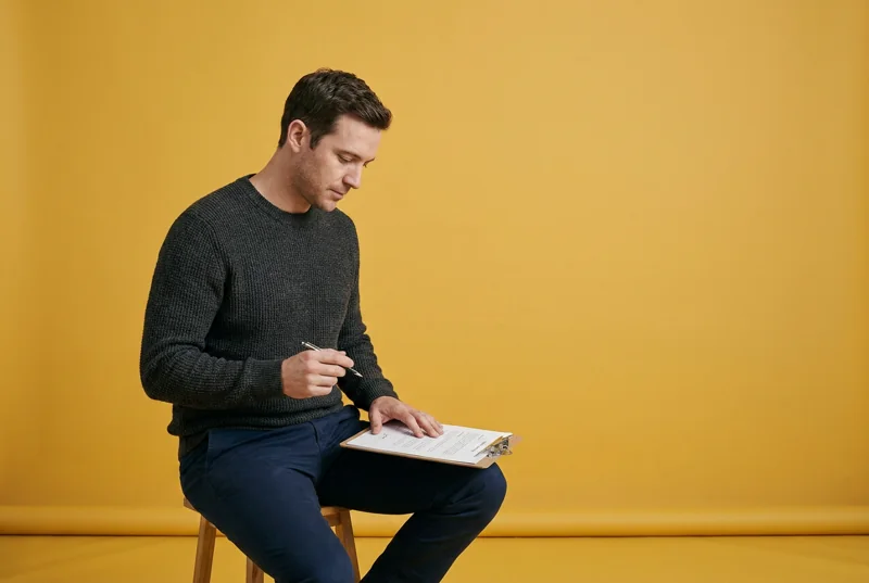 A man seated on a stool with a clipboard on his knee, pen paused above the page before signing, in front of a yellow backdrop.