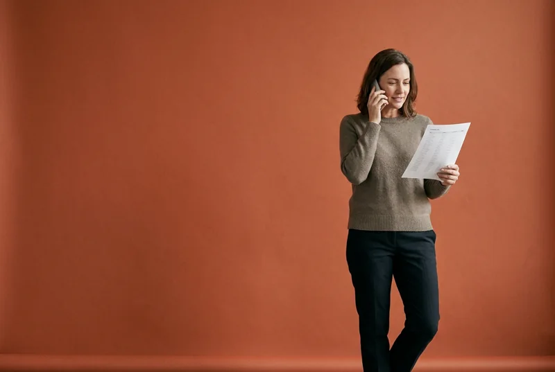 A woman on the phone holding a printed customer list, mid-conversation, in front of a terracotta backdrop.