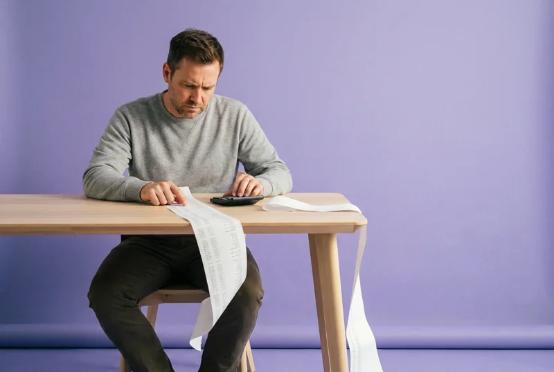 A man at a timber table tracing a finger down a long paper printout that spills over the table edge, in front of a lilac backdrop.